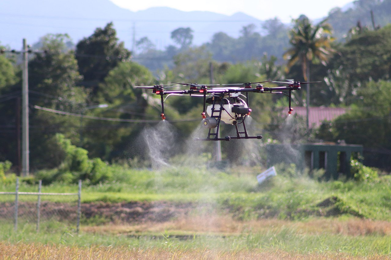 A multirotor agricultural spray drone hovering low over a field with its nozzles actively spraying