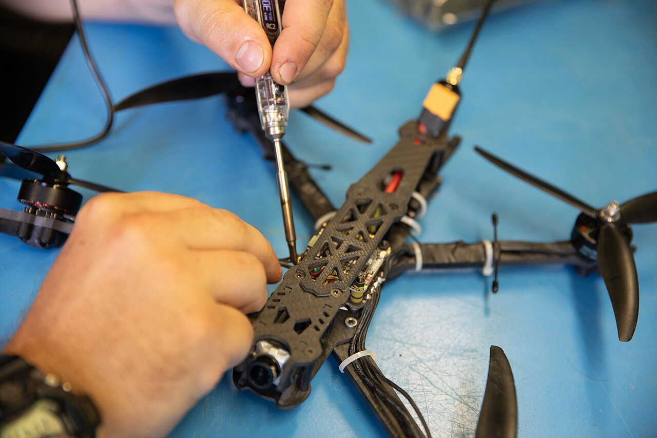 Hands soldering wiring on a carbon-fibre FPV drone frame on a blue workbench mat — the practical reality of AI drone integration