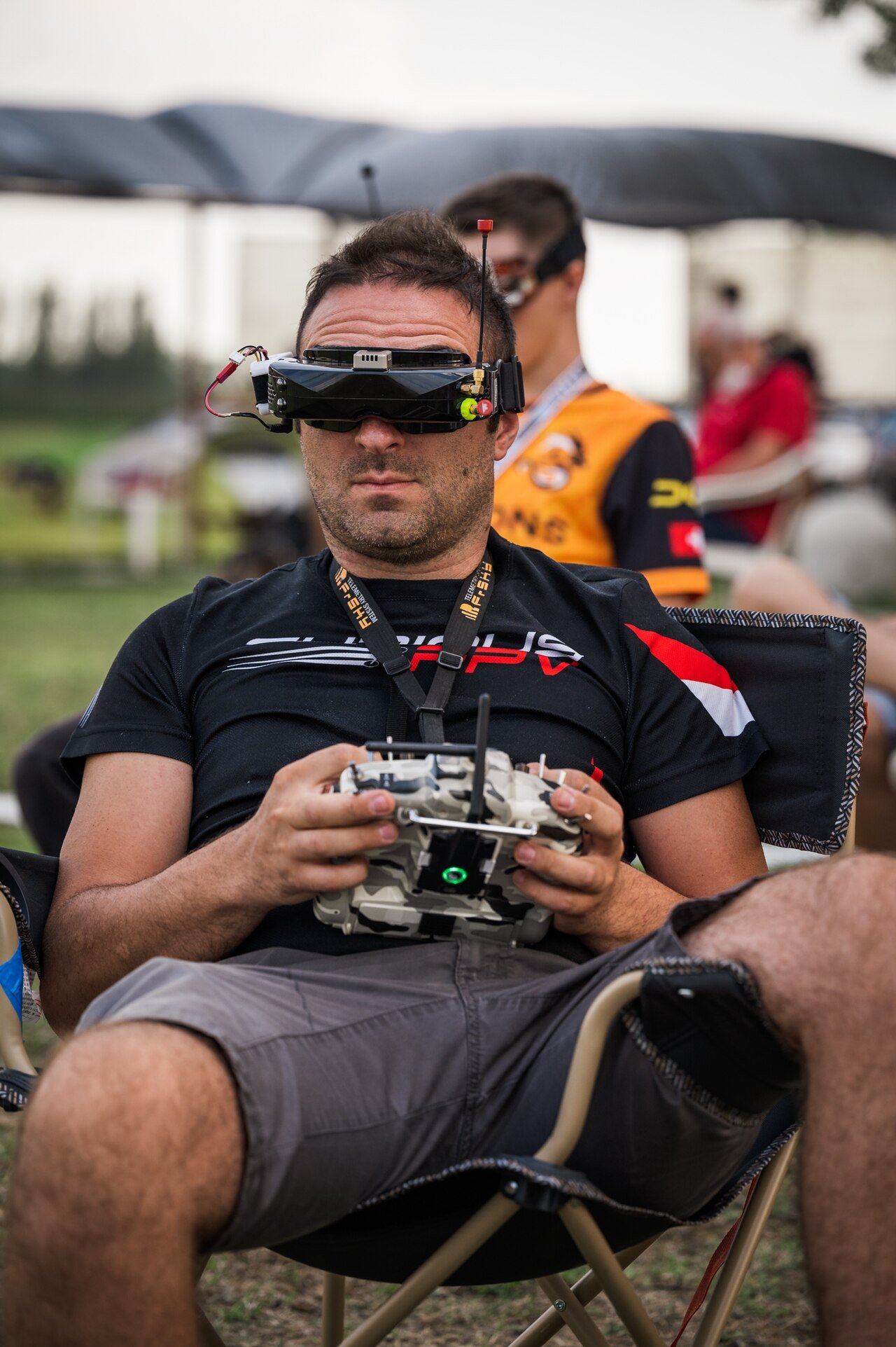 An FPV pilot wearing video goggles and holding a radio transmitter, flying a first-person-view drone at an outdoor event