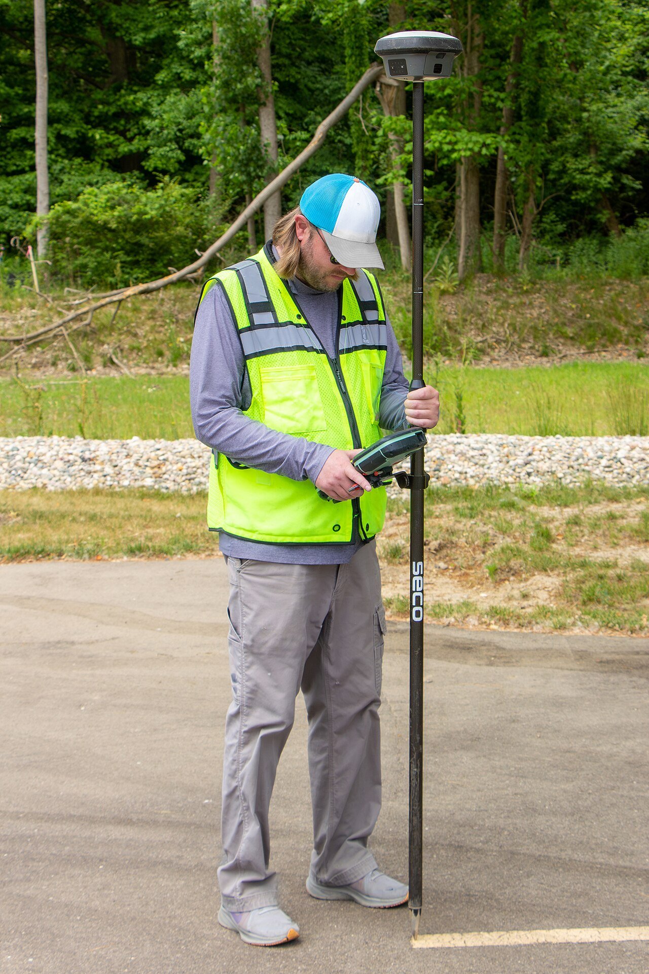A surveyor holding an RTK GNSS rover pole with the receiver antenna on top and a handheld controller, working on a paved area